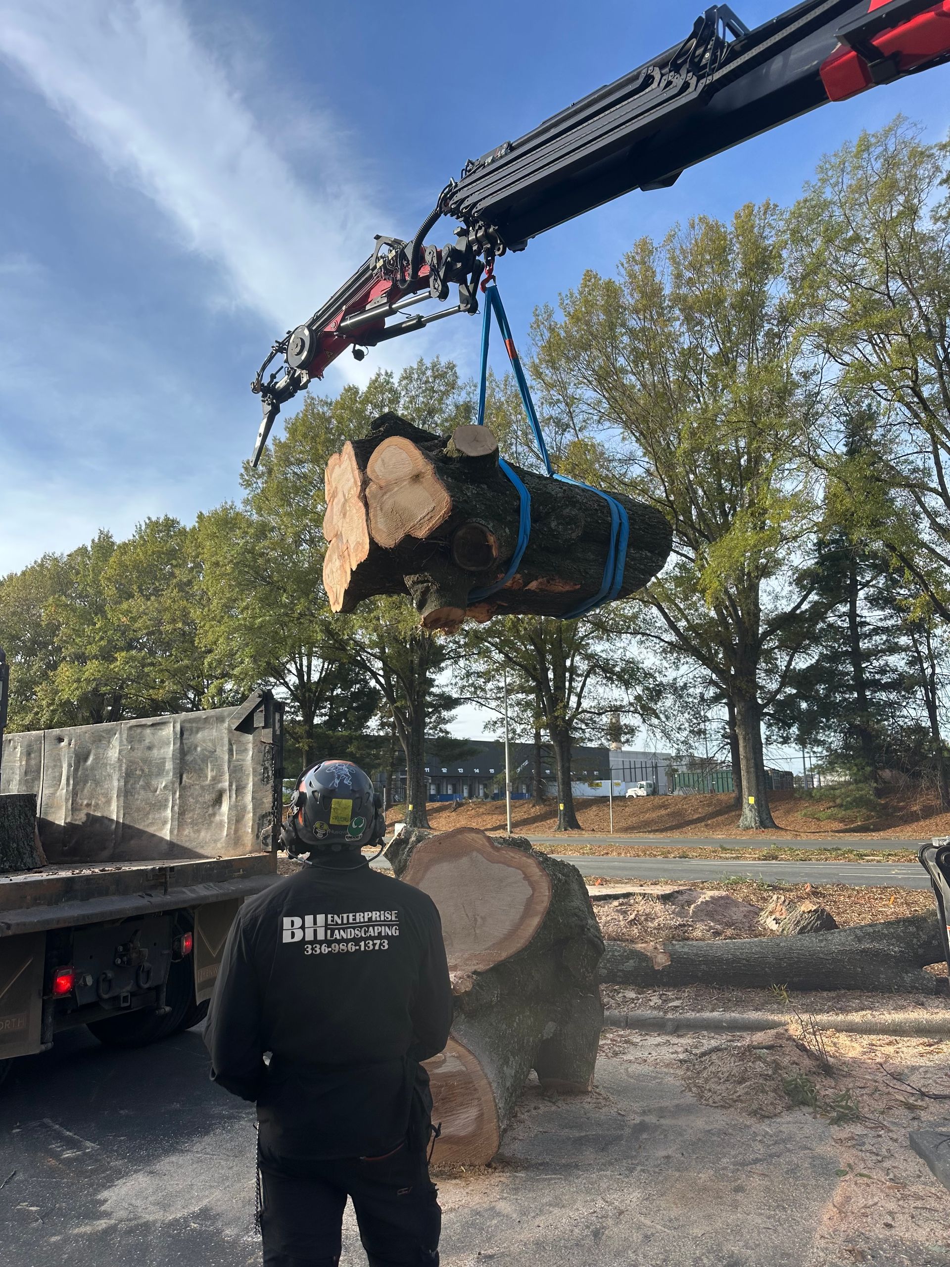 A man wearing a shirt that says gnf is standing in front of a large log being lifted by a crane
