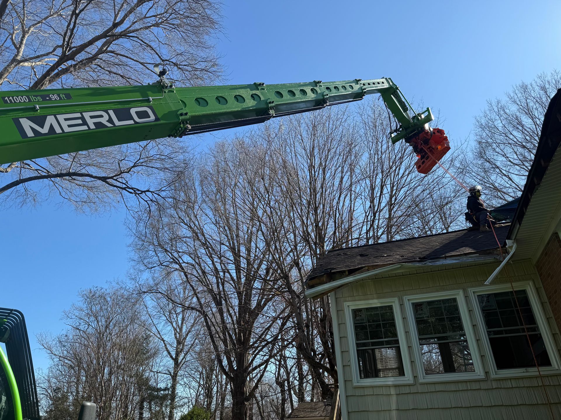 Green Merlo telehandler trimming a tree branch near a house with windows under a blue sky.