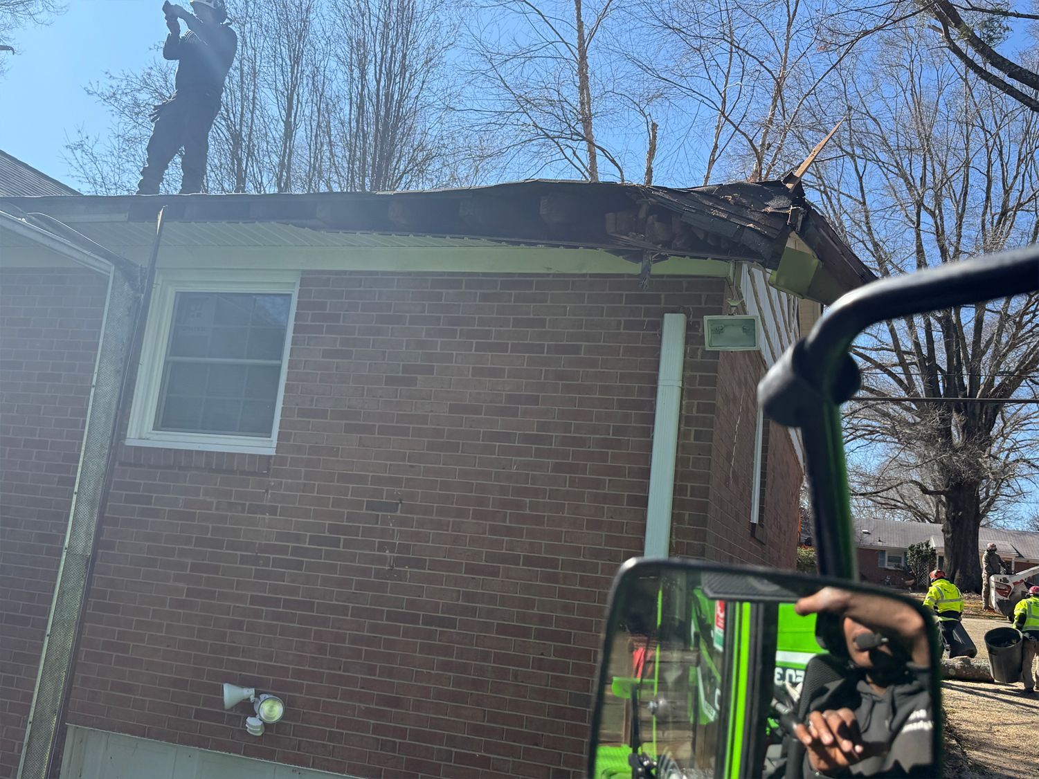 Man on a roof repairs a damaged section, other workers in the yard. Brick building, trees, clear sky.