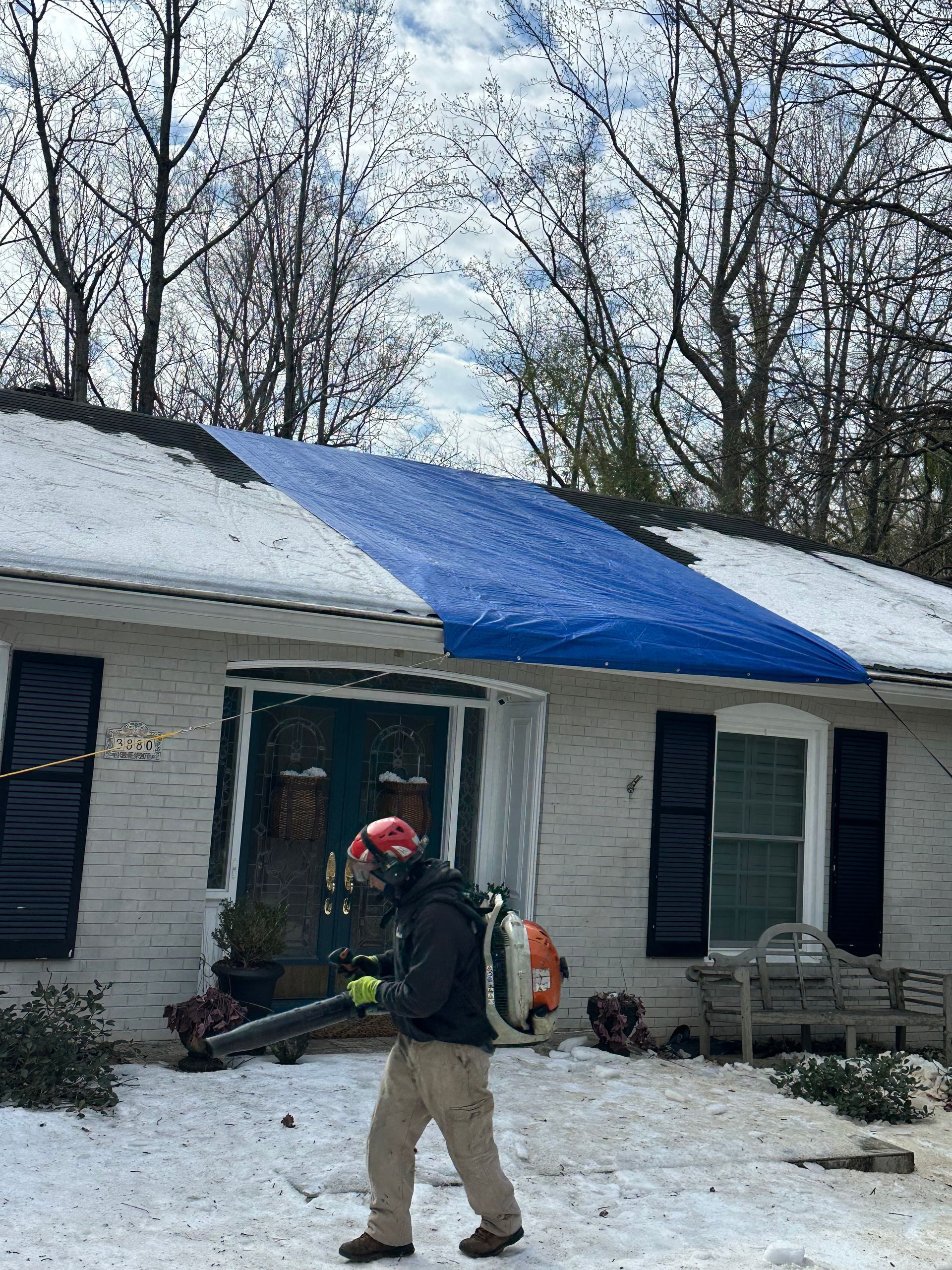 Person with leaf blower clearing snow from a roof covered with a blue tarp.