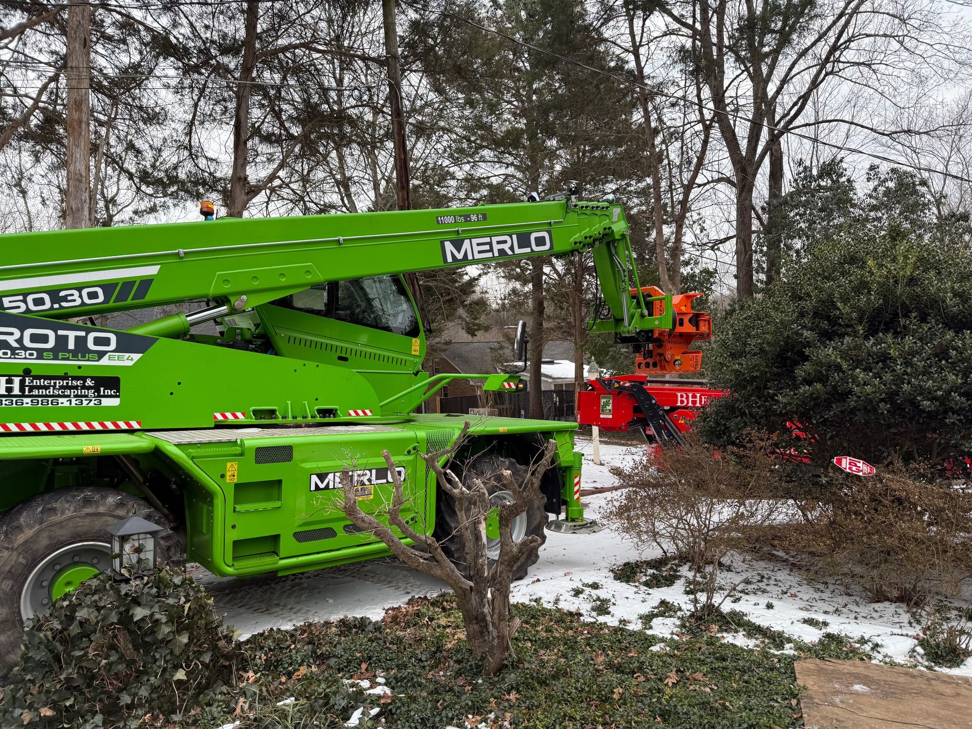 Green Merlo telehandler operating near a red industrial machine, shrubs, and snowy trees.