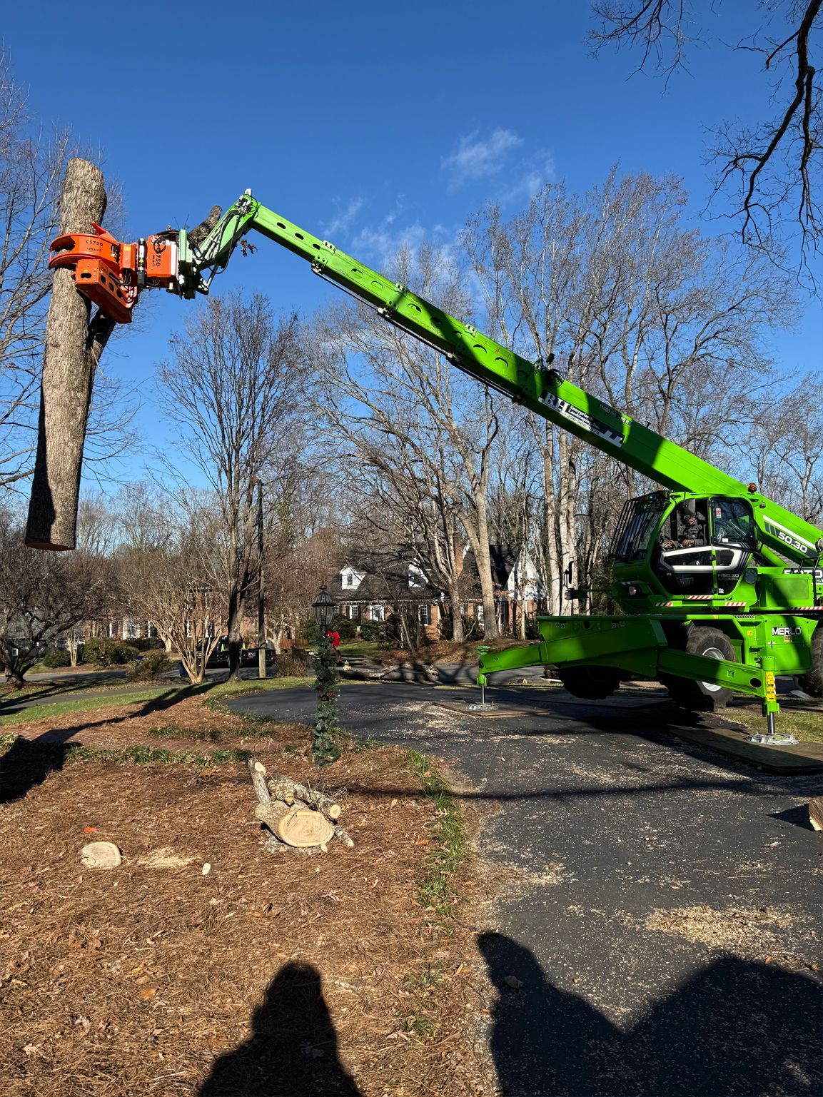 Green tree removal machine cutting a tree trunk on a paved driveway; clear blue sky.