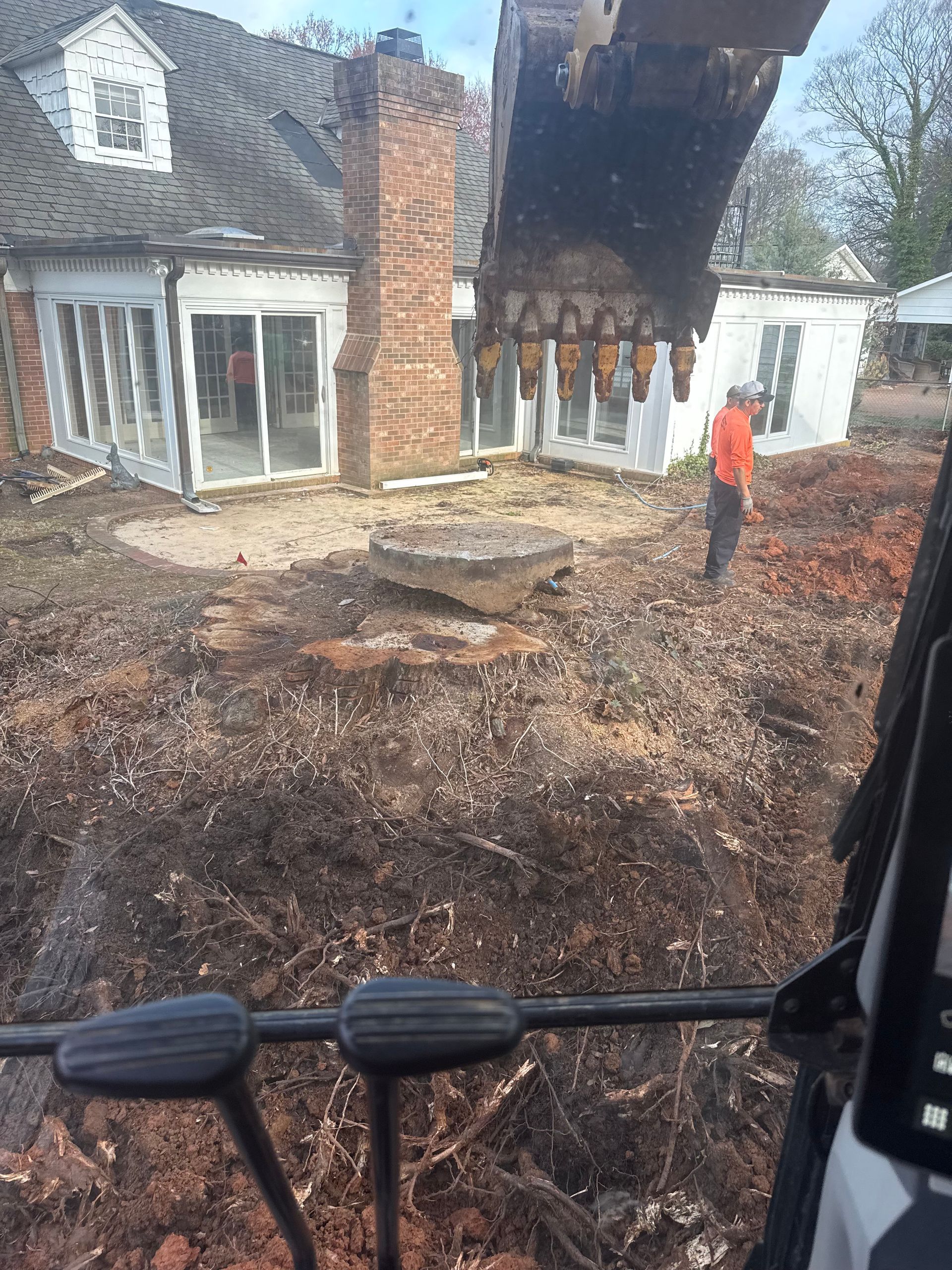 Excavator removing earth near a house with a brick chimney. A worker in an orange shirt watches.