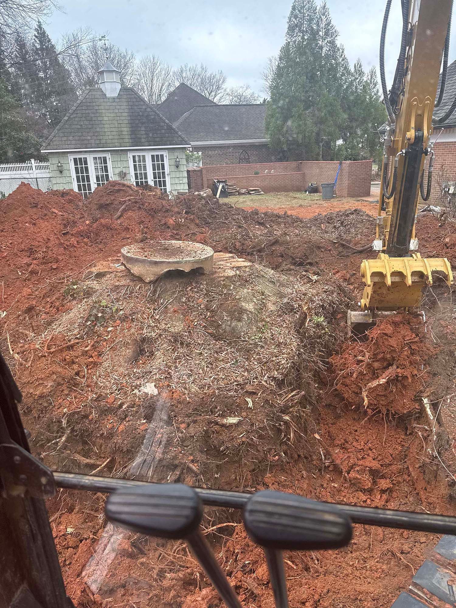 An excavator digs in a yard; red soil, tree stump, and building in the background.