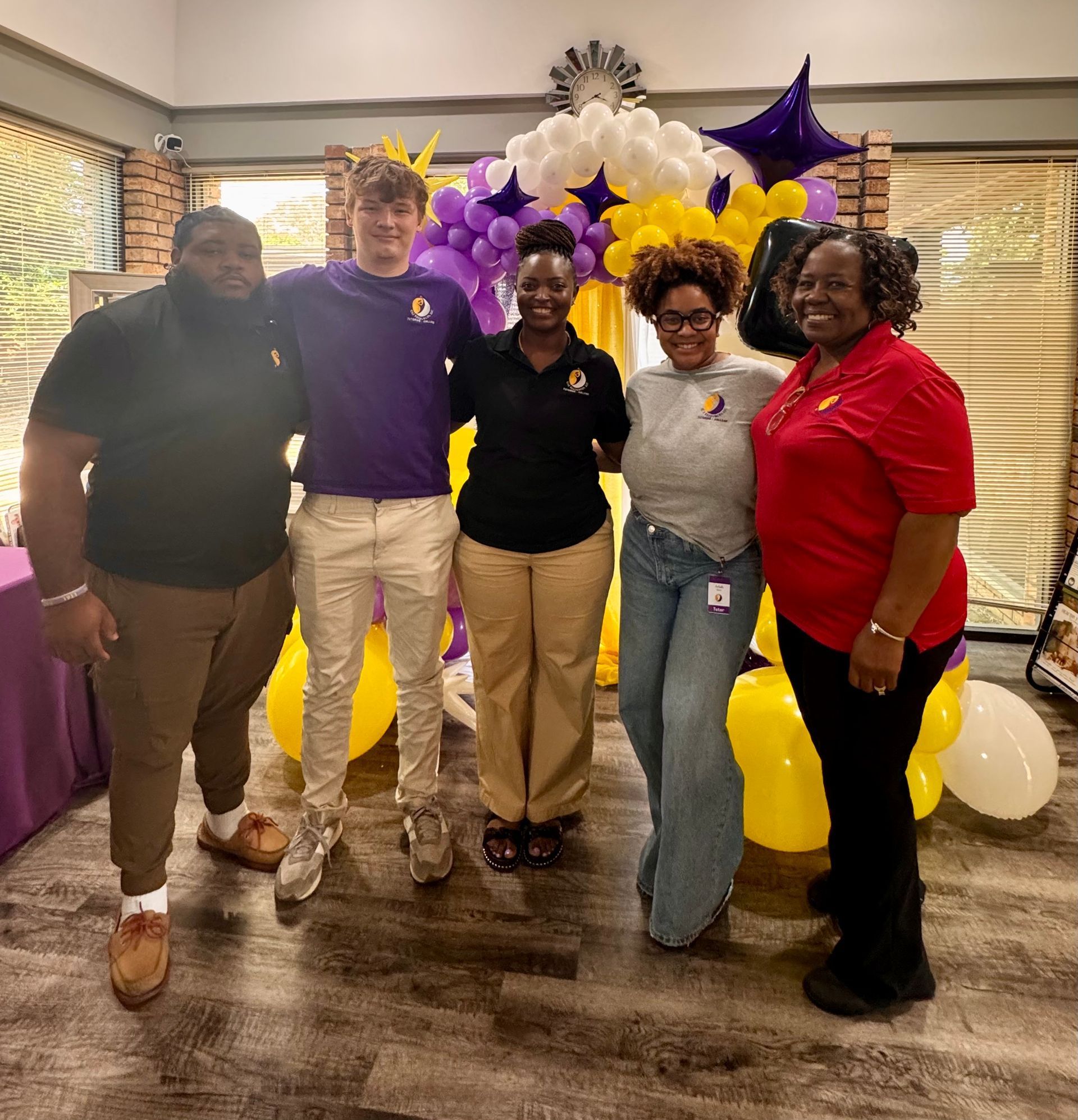 Group of people smiling with purple and gold balloons indoors.