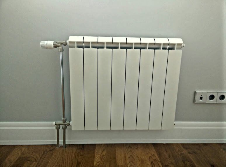 White radiator on a light gray wall above wooden flooring, with chrome pipes and a temperature control knob.