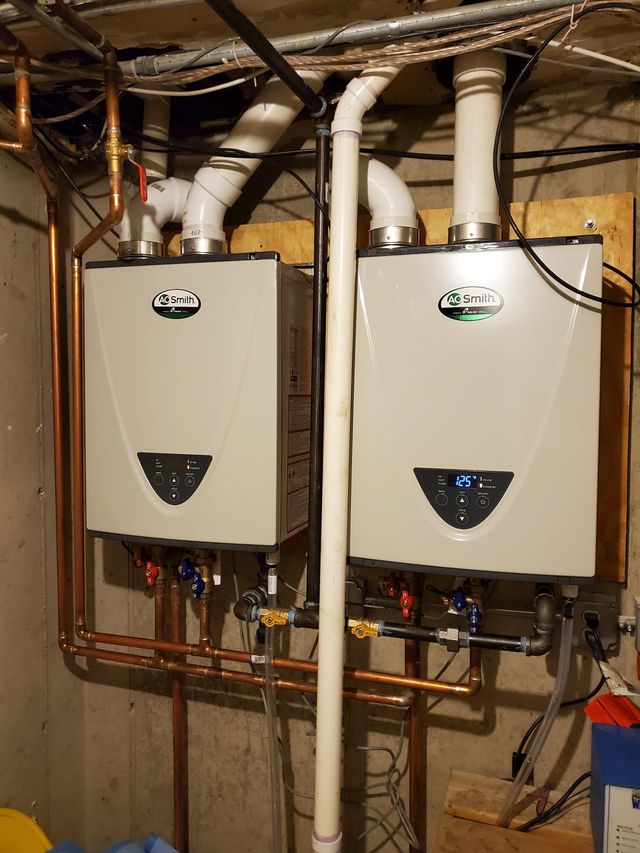 Two wall-mounted, white tankless water heaters with copper piping against a concrete wall.
