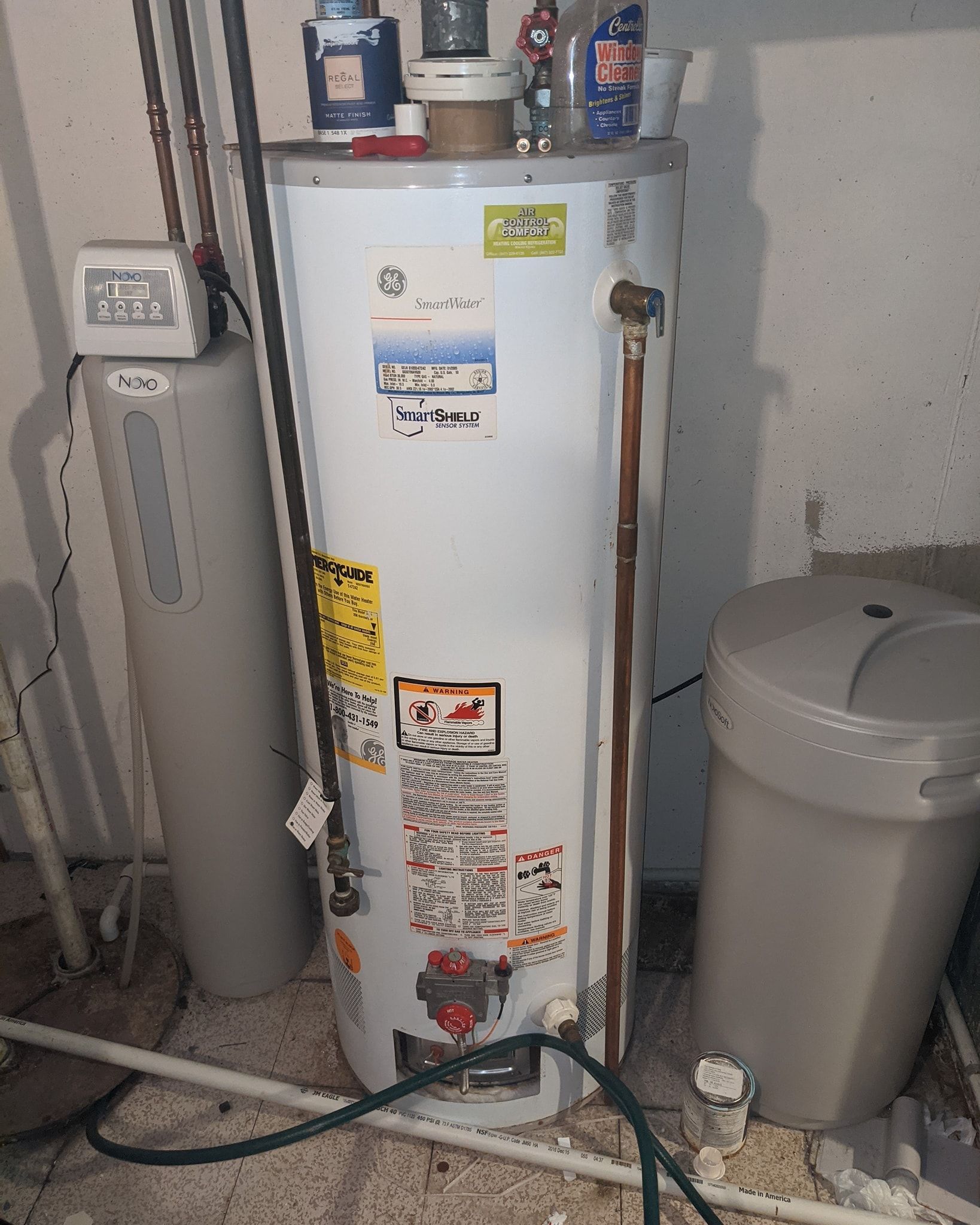 Water heater and water softener in a utility room. White and gray appliances, pipes, and a trash can.