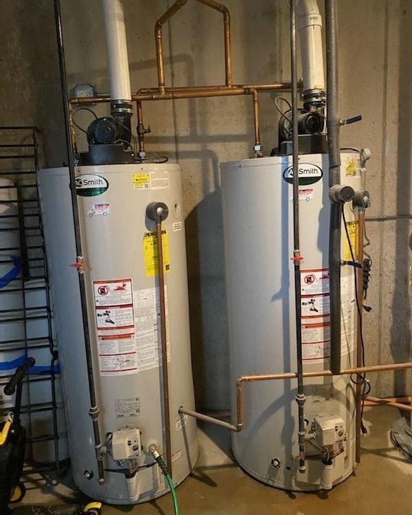 Two tall, gray water heaters in a basement, connected by copper pipes and white vent pipes.