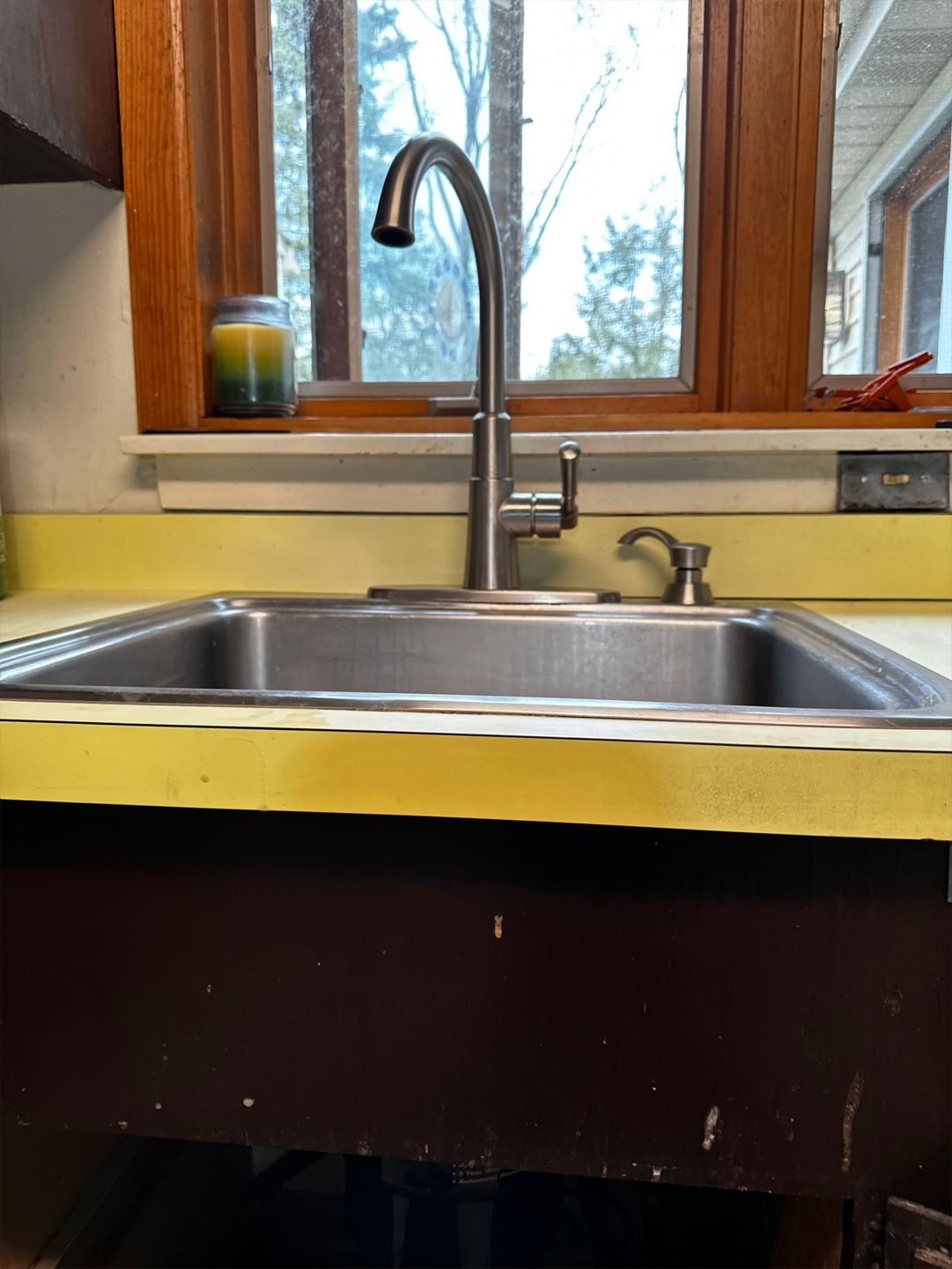 Stainless steel kitchen sink with faucet, yellow counter, dark brown cabinet, window in background.