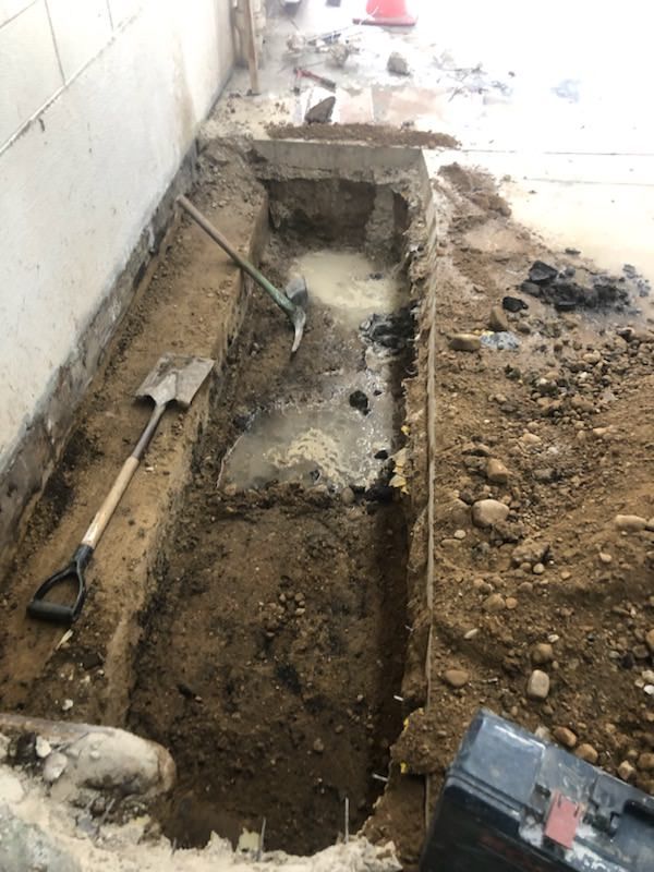 Trench dug alongside a building, containing tools and standing water. Dirt and concrete visible.