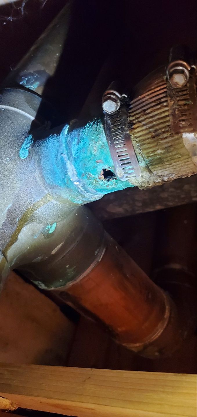 Leaking copper water pipe with blue corrosion and a clamp, in a dark wooden space.