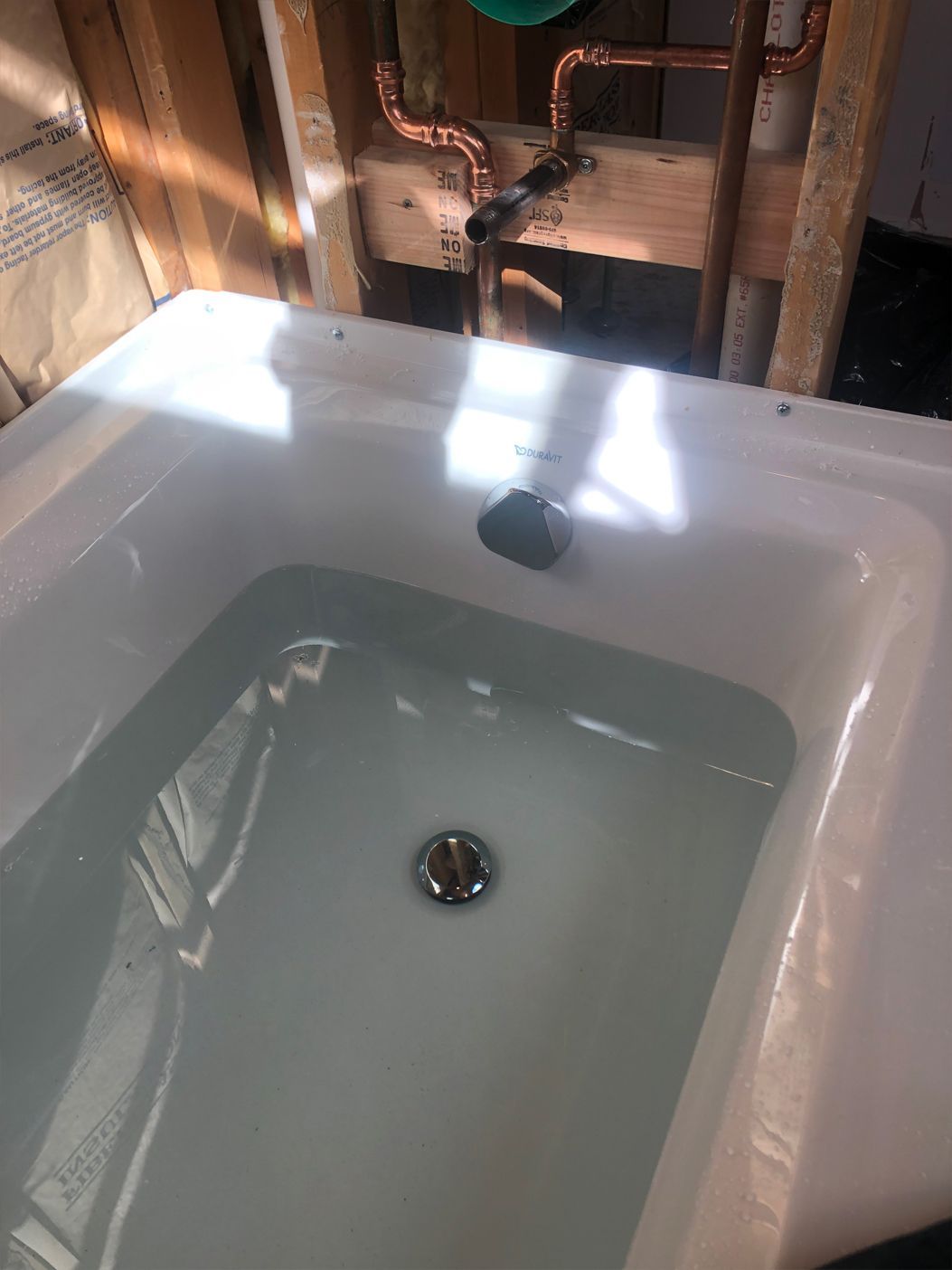 Bathtub filled with water, drain visible. Copper pipes and wood studs in background.