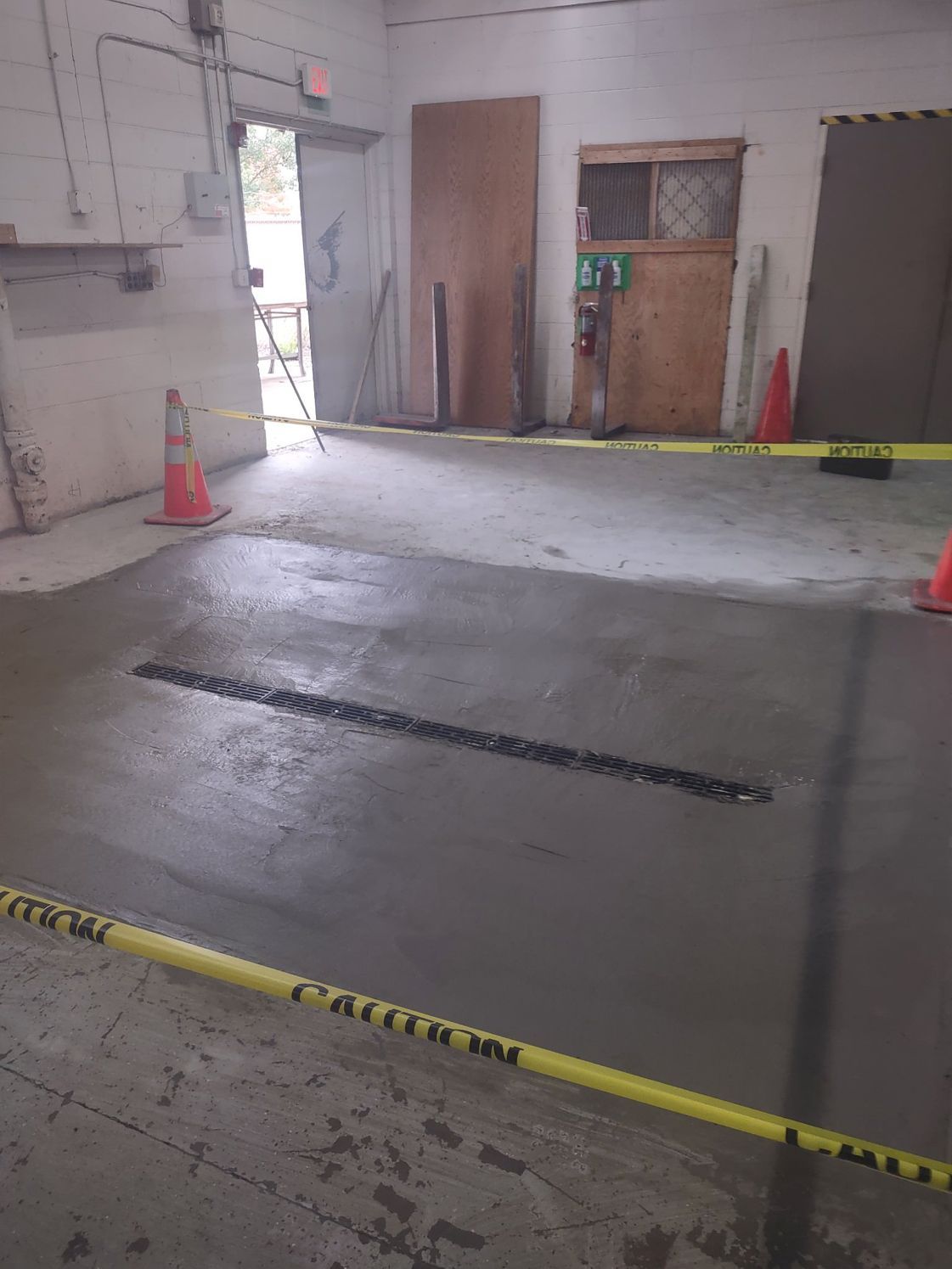 Freshly poured concrete floor with drainage grate, cordoned off with caution tape and cones.
