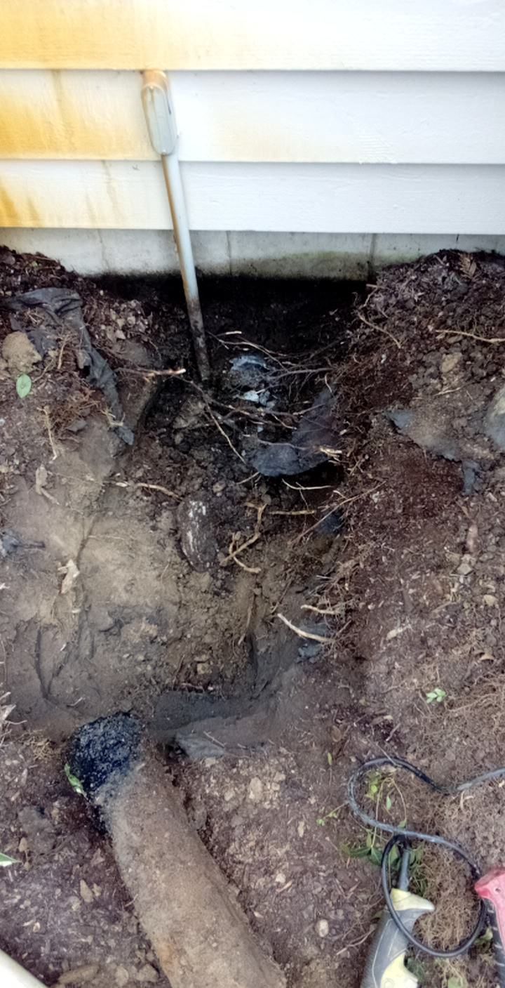A small trench by a house foundation with a pipe, exposed soil and mulch.