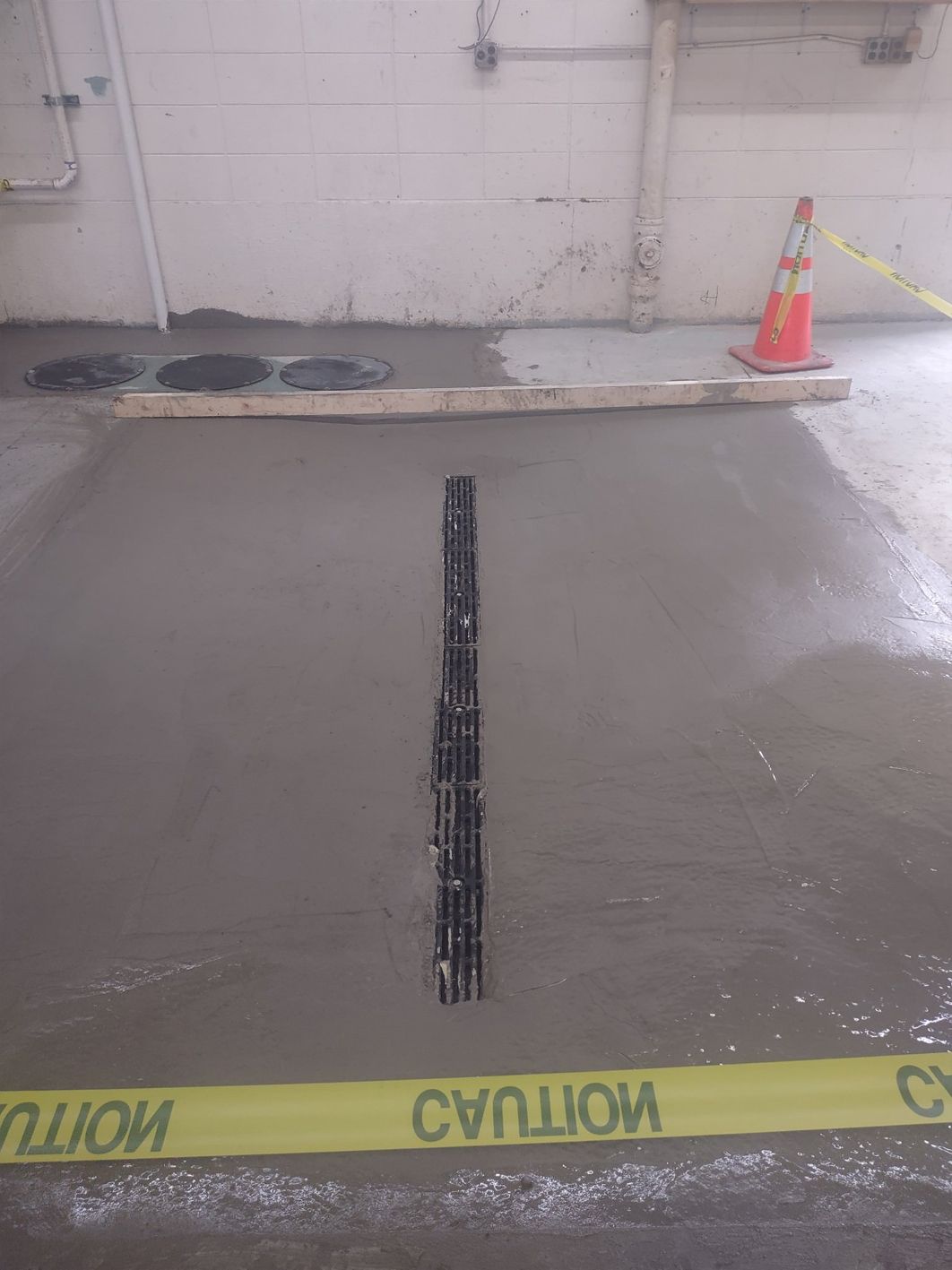 Fresh concrete floor with black grate in the center, caution tape, and an orange cone.