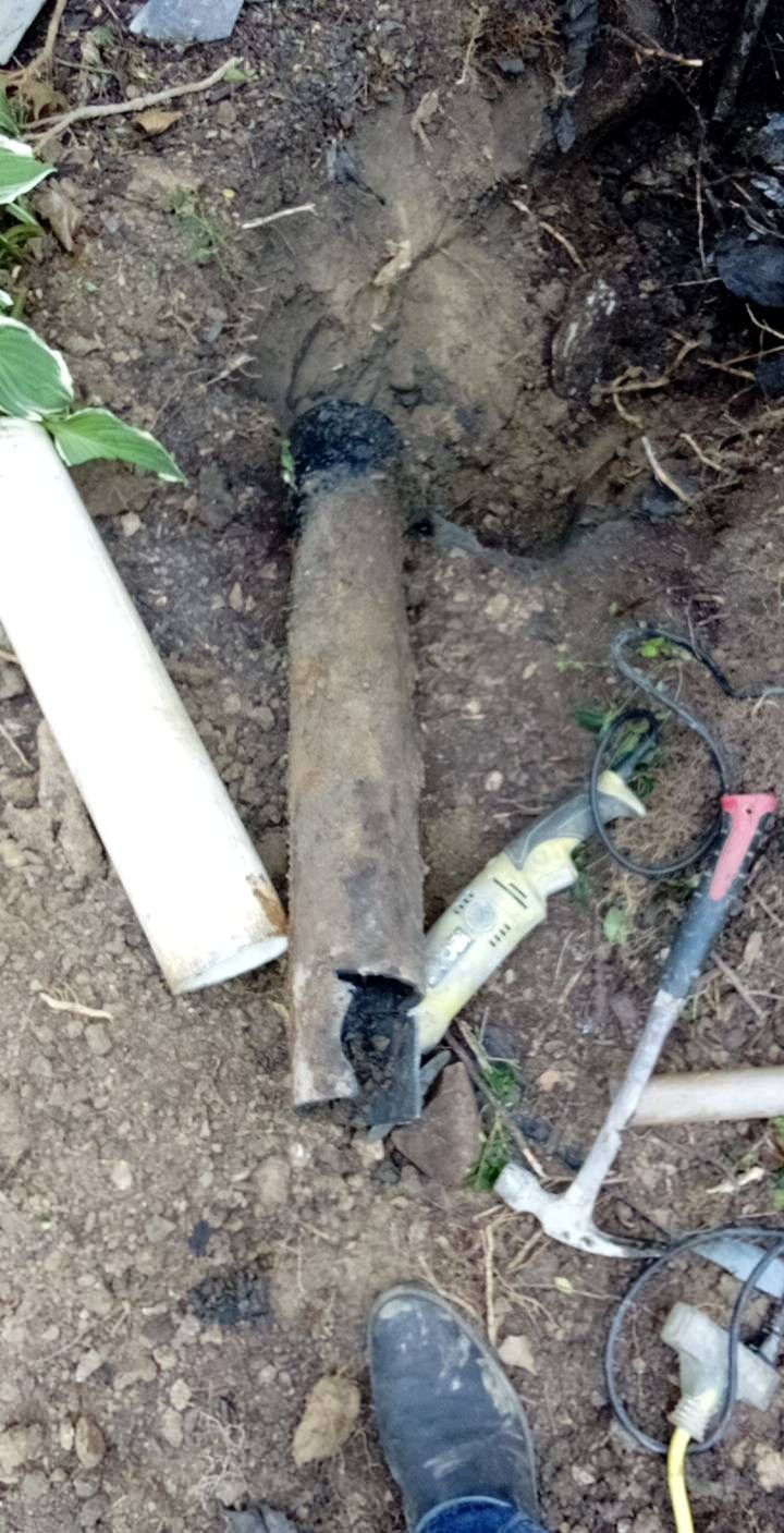 A cylindrical, charred object is partially buried in dirt. A white pipe and tools lay nearby.