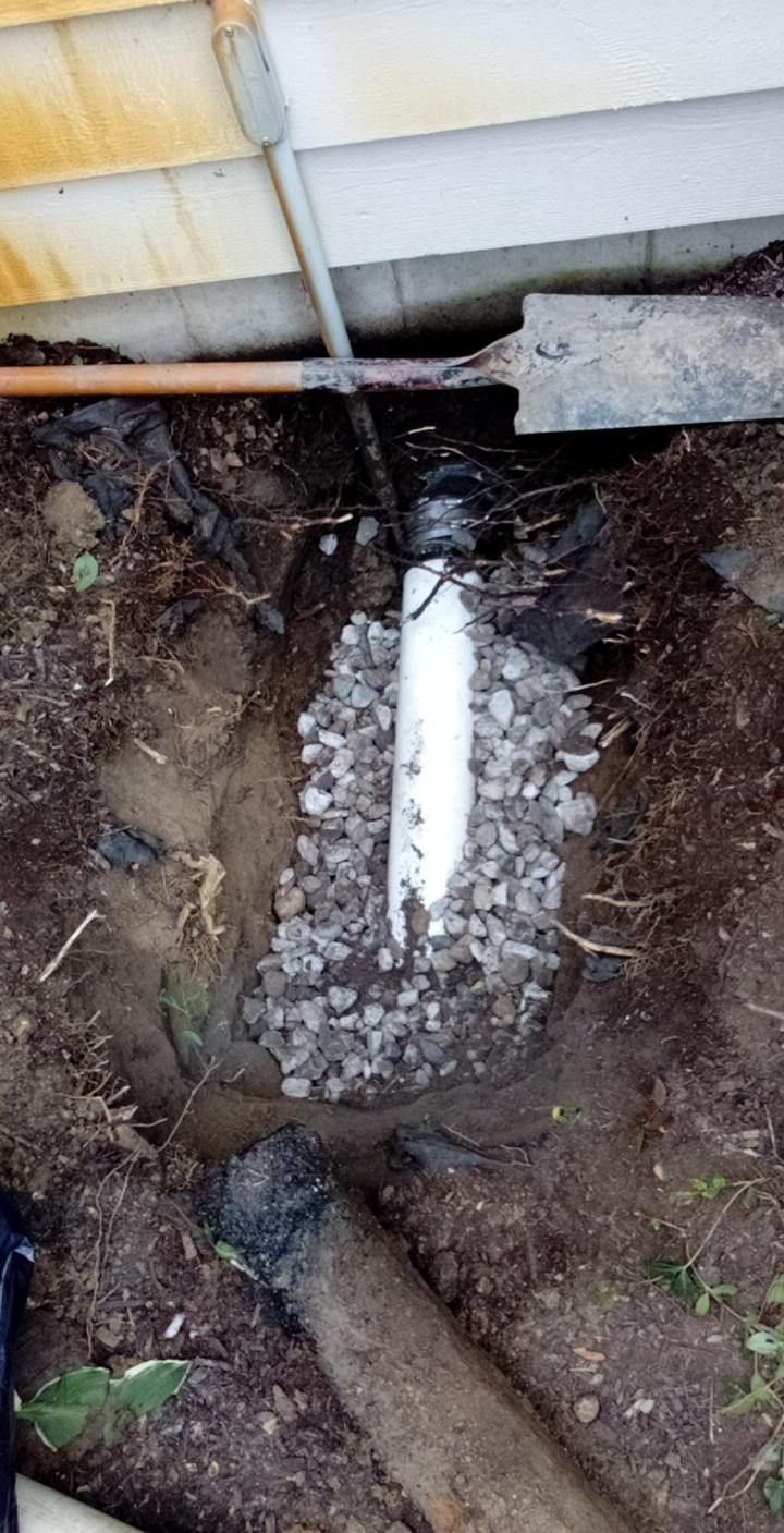 White PVC pipe in gravel-filled trench. Dark drain pipe connects to it near a building's foundation.