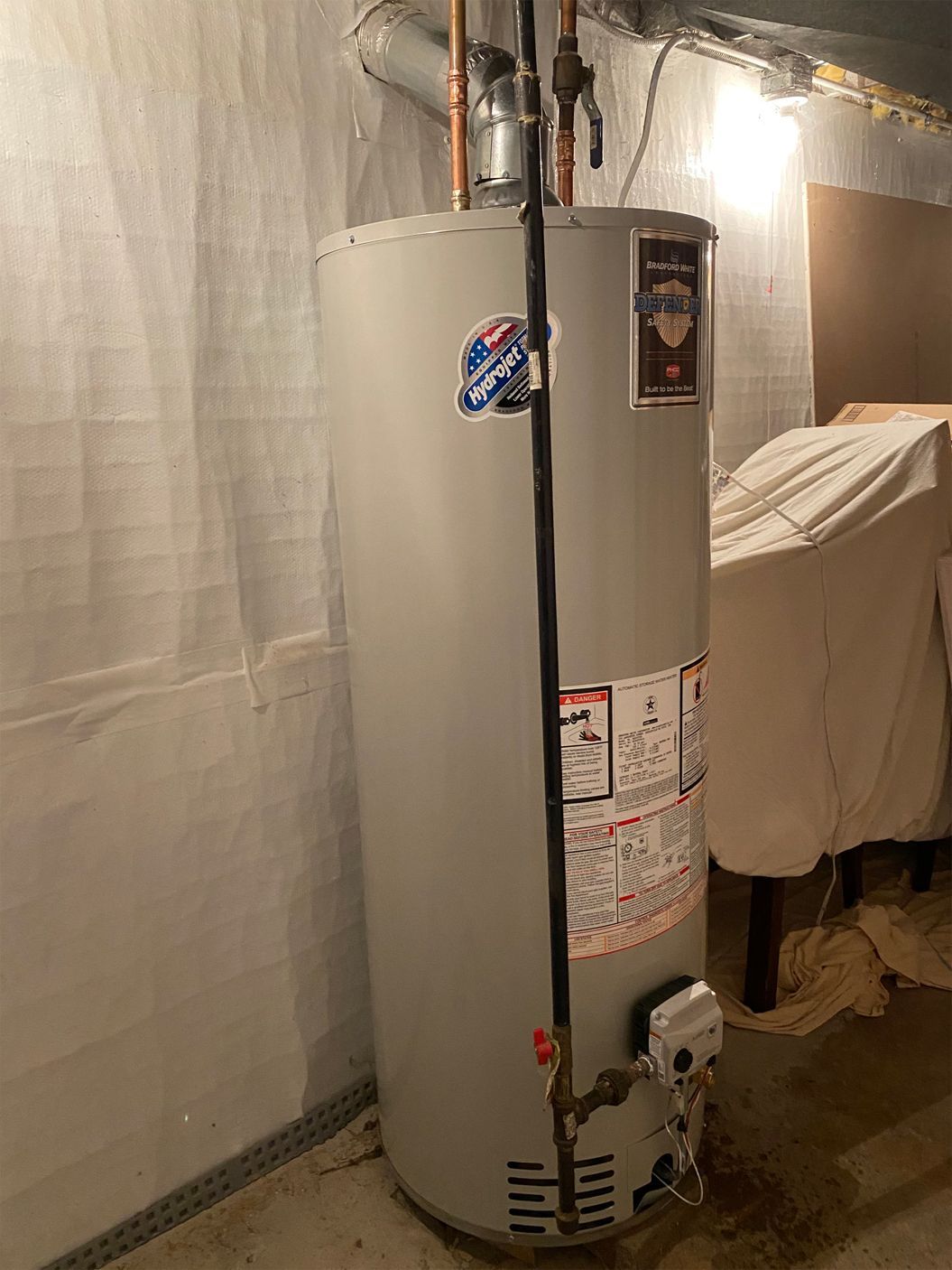 A tall, cylindrical water heater in a basement.  It has copper pipes and a black gas line.