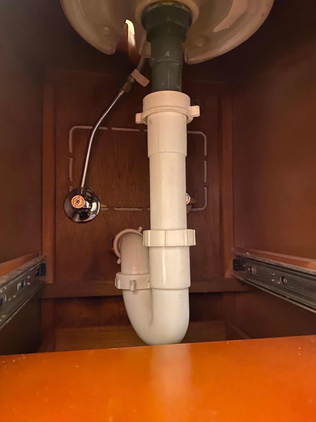 Plumbing under a sink, showing pipes, drain, and a chrome-plated ball float valve. Brown cabinet interior.