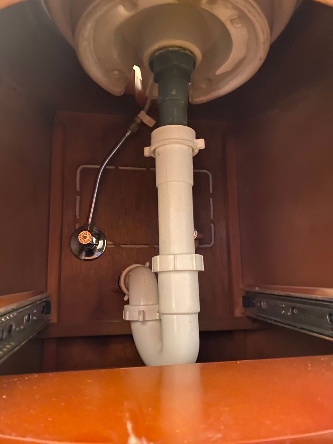 Under-sink plumbing: white pipes, P-trap, drain line, and pop-up drain assembly, within a wooden cabinet.