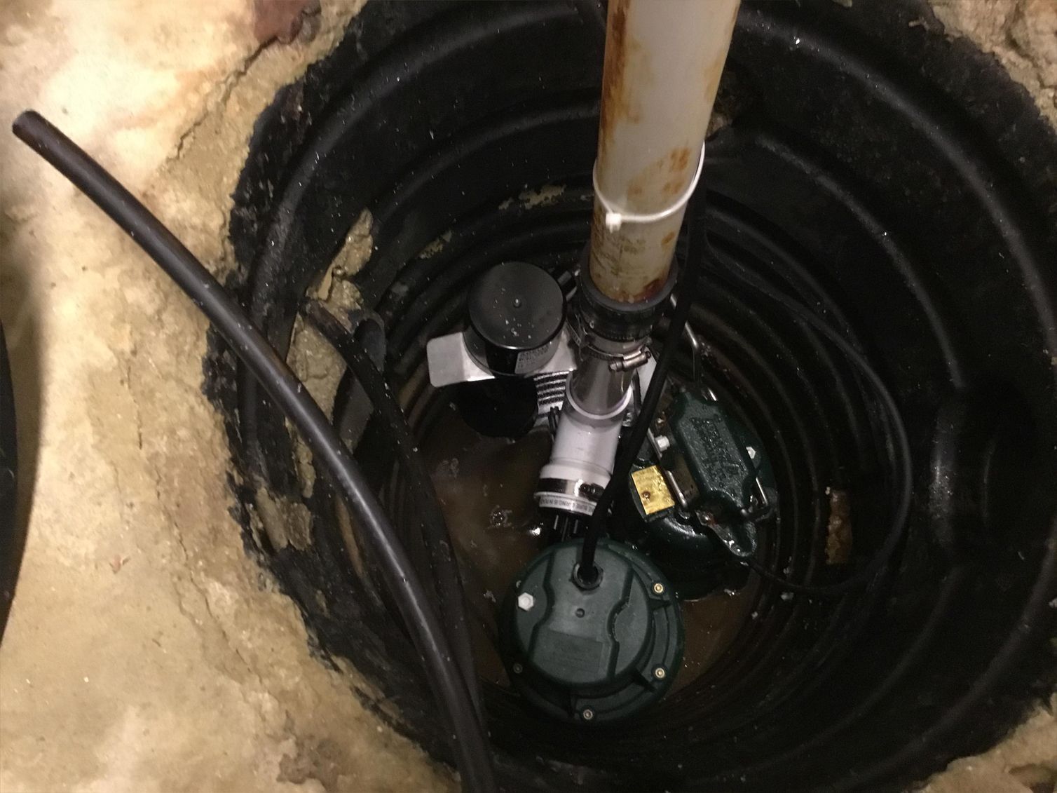 Sump pump in a black basin, with pipes and electrical components in a concrete setting.