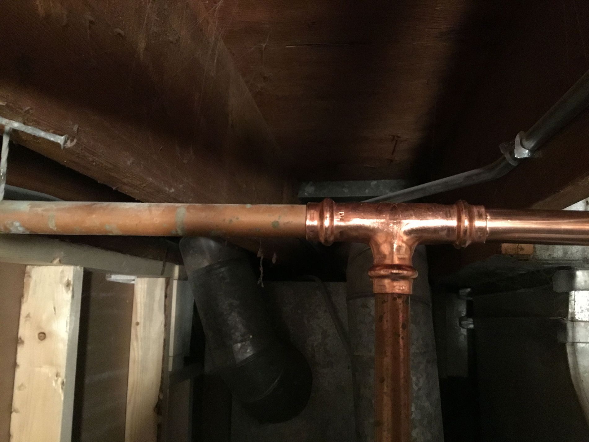 Copper plumbing pipes in a basement, including a T-joint, run horizontally and vertically against a wooden structure.