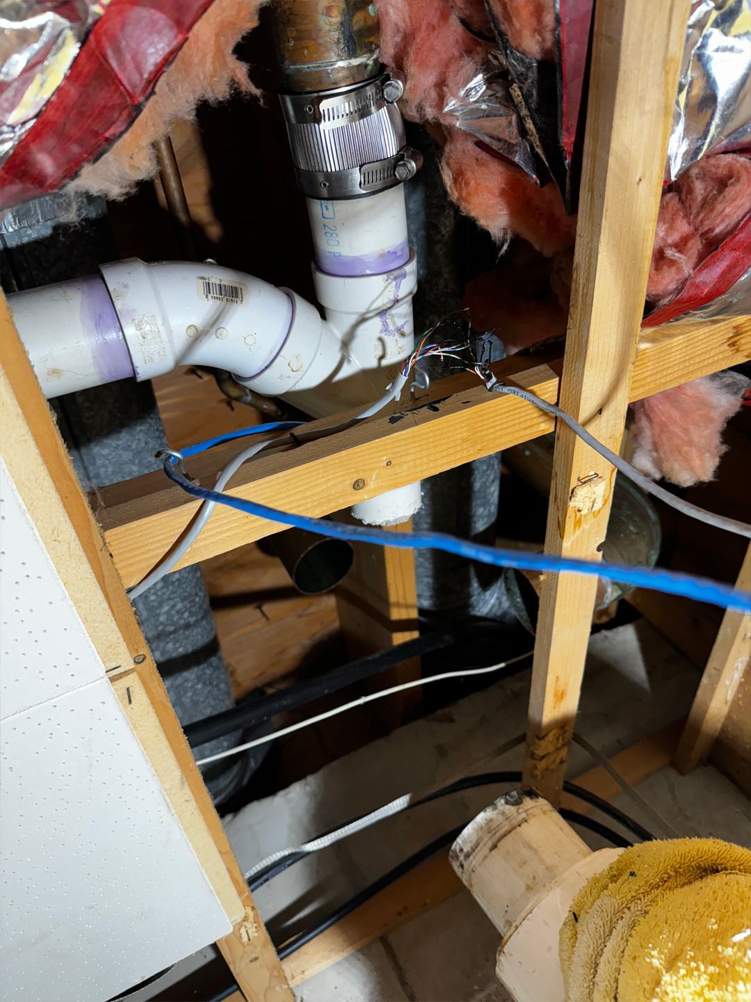 Pipes and plumbing fixtures within a wood-framed wall, showing white and purple PVC pipes connected with clamps, wiring.
