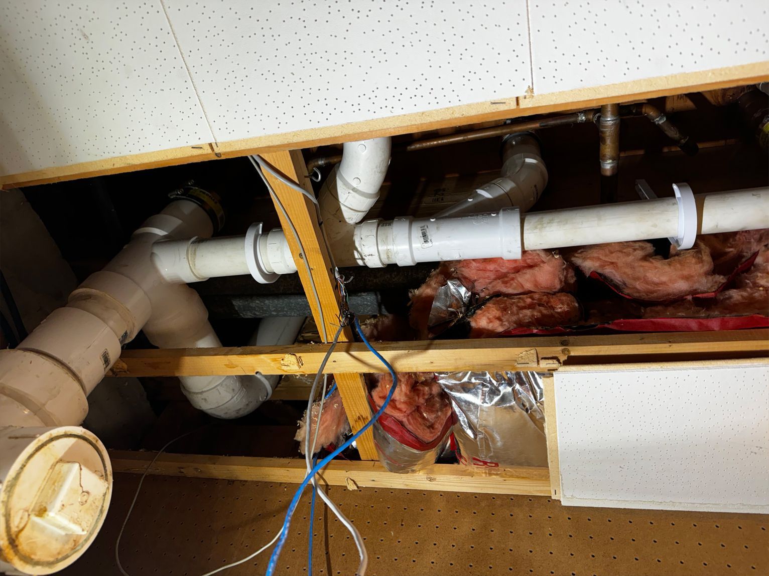 Pipes and plumbing fixtures exposed inside a ceiling, with wooden framing, insulation, and electrical wires visible.