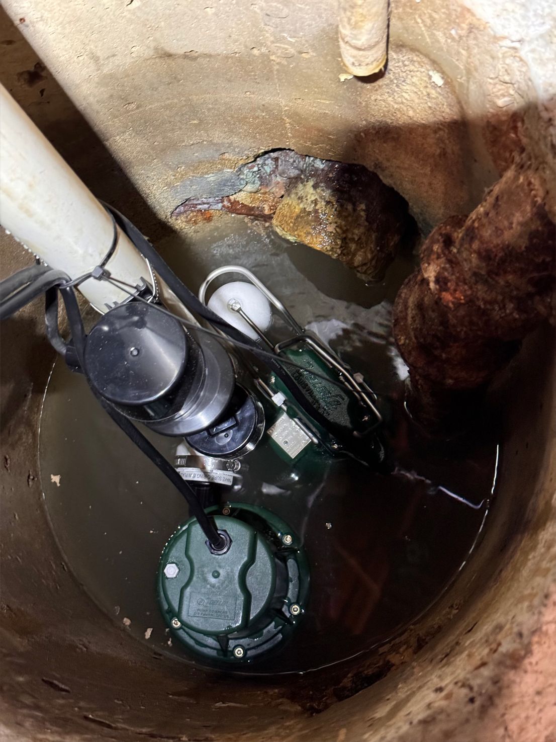 A sump pump in a concrete pit. Dark water surrounds the pump, with a white pipe and rust-colored pipe visible.