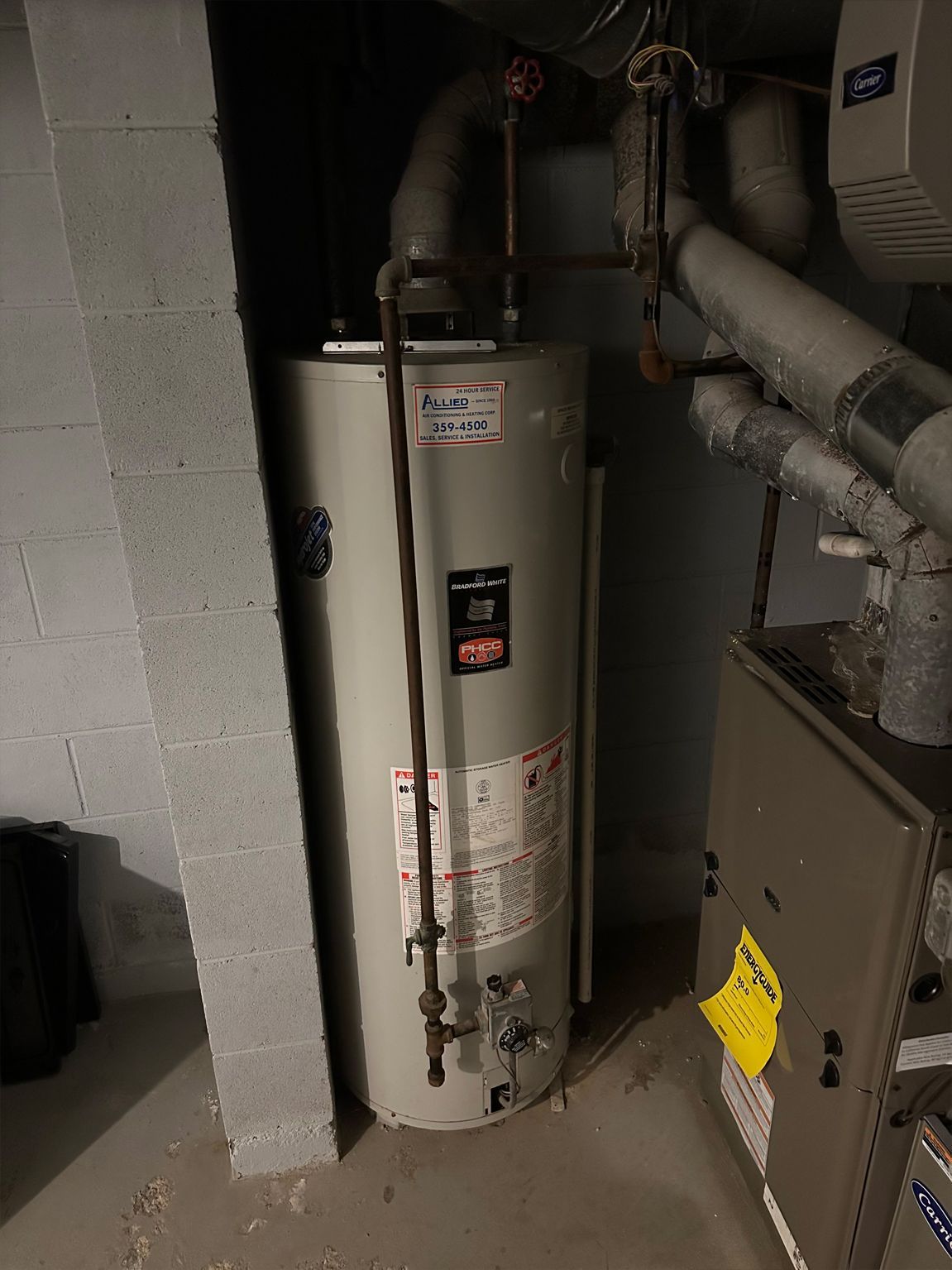 A tall, cylindrical water heater in a basement setting. Pipes and vent near a furnace. Gray and metal.