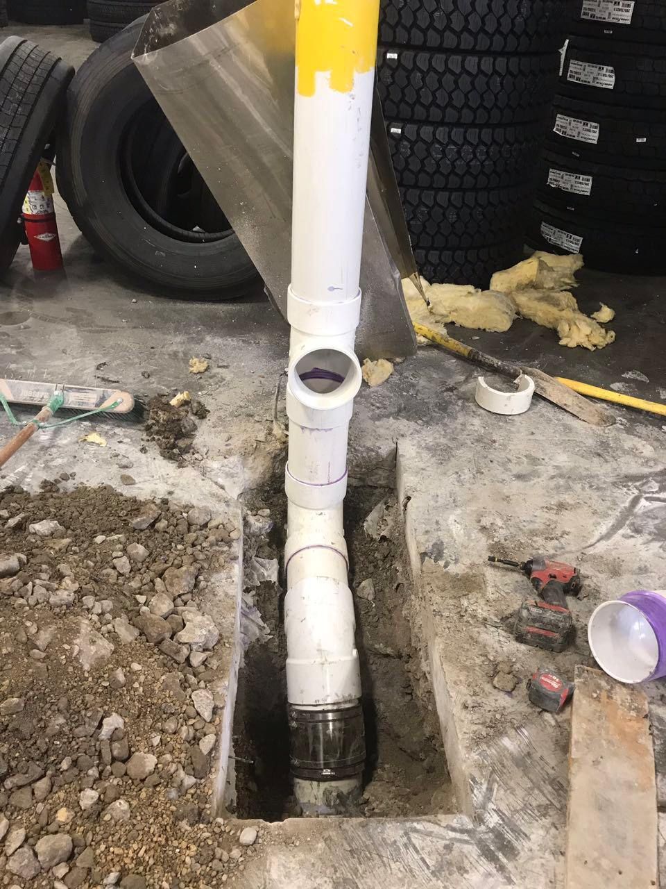 White PVC pipe and fittings installed in a concrete trench, with a yellow-painted top section; tires in background.