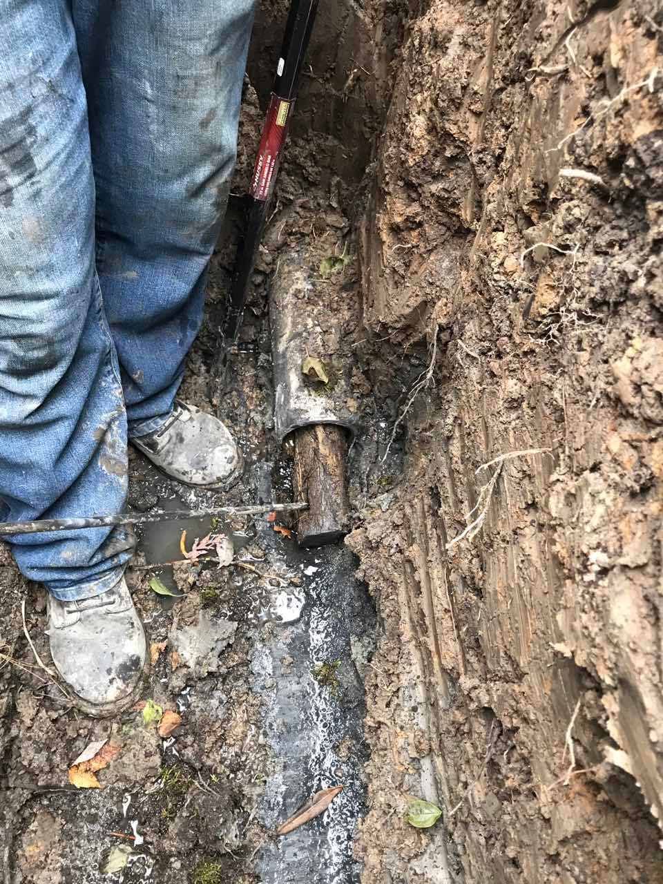 Person in muddy trench working on a pipe. Visible are the person's legs, boots, and tools, surrounding the pipe.