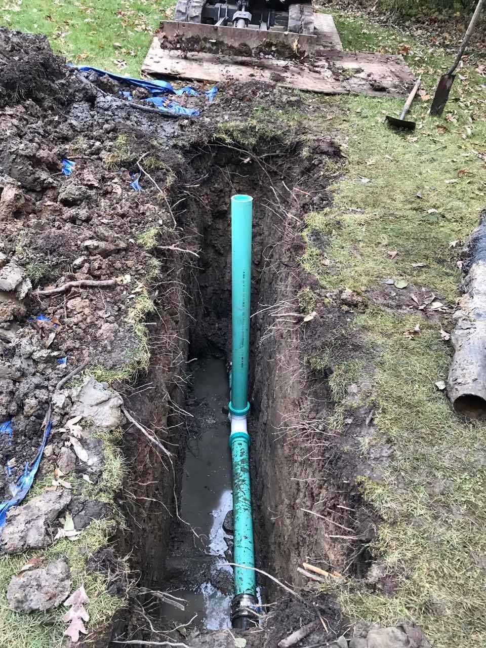 Trench dug in yard with a green pipe visible, water at the bottom, and a shovel nearby.