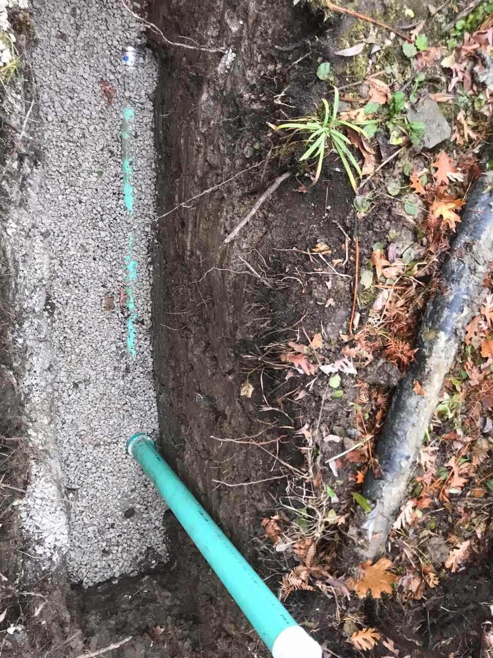 Trench with gravel and green pipe; dark soil, leaves, and a gray pipe nearby.