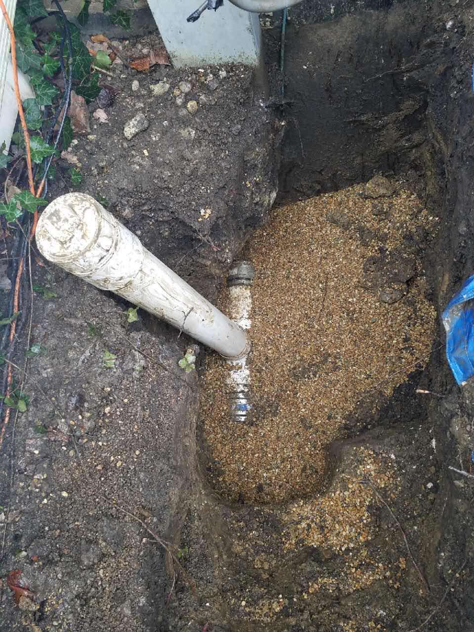 White PVC pipe connected to a clear plastic fitting buried in dirt and sand in a hole.