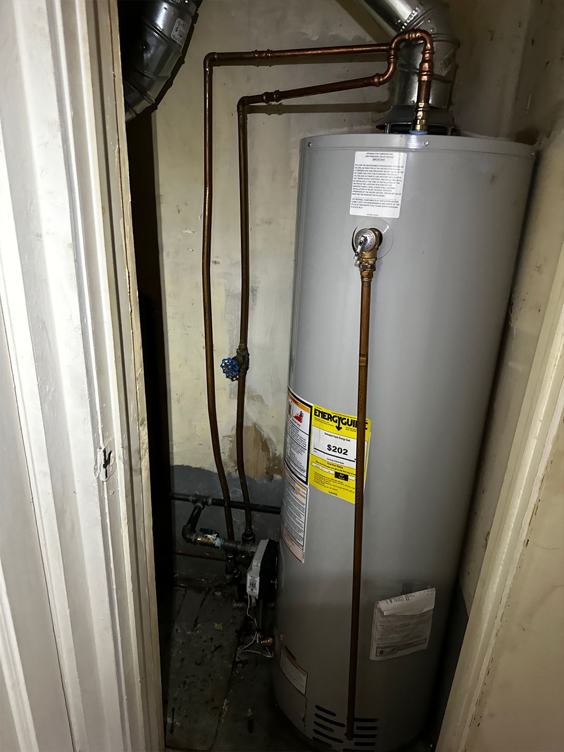 Water heater in a cramped, enclosed space, with copper pipes and a metal vent.