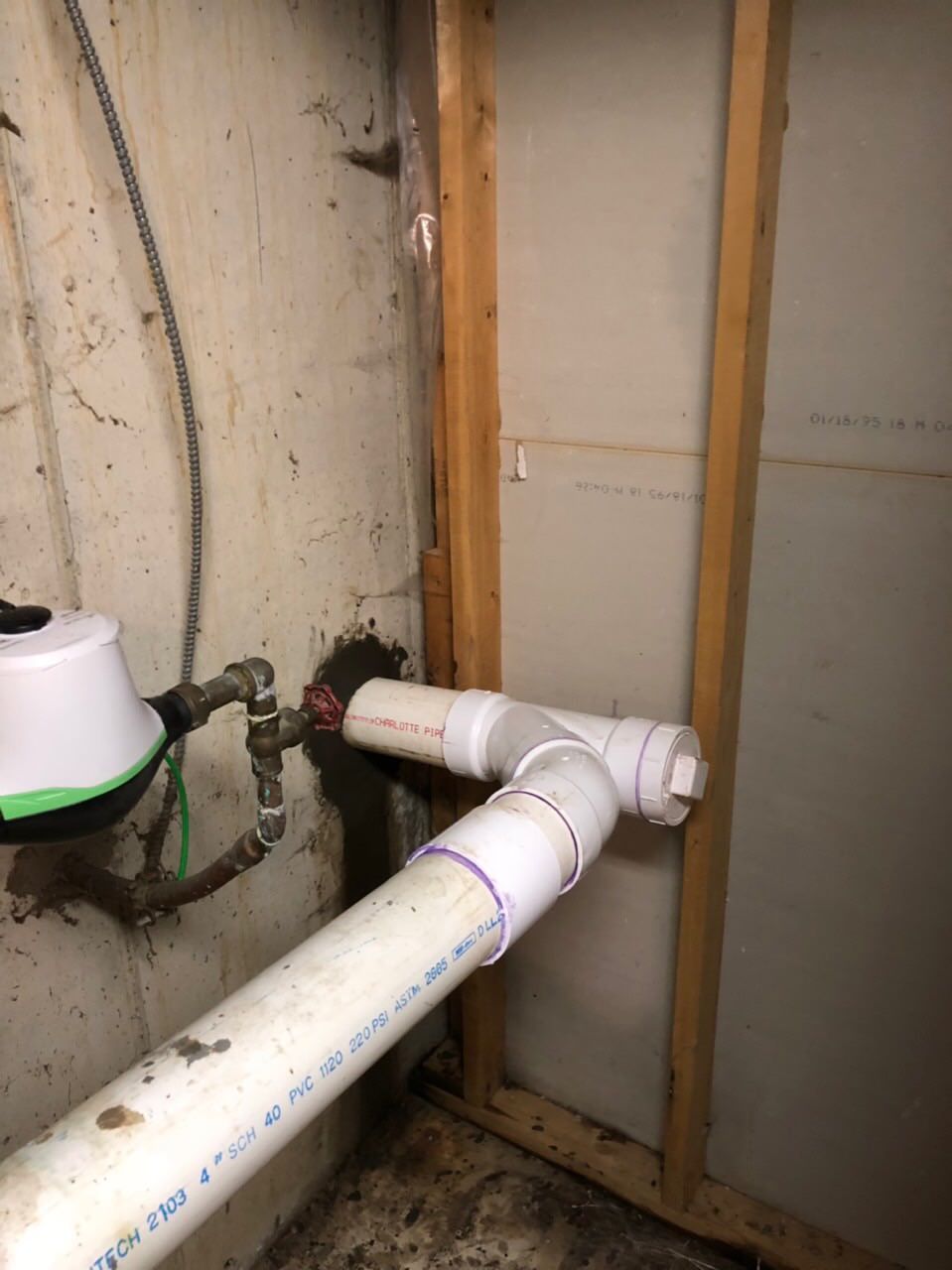 Plumbing pipes with joints and fittings in a corner of a basement. White pipes against a light brown wall.