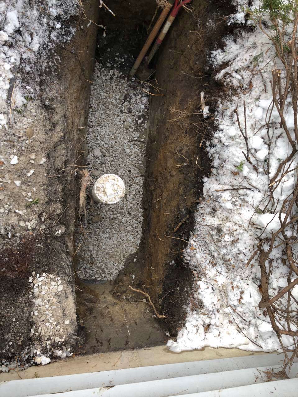 Trench dug in the ground, with a white cylinder and gravel, surrounded by dirt and snow.