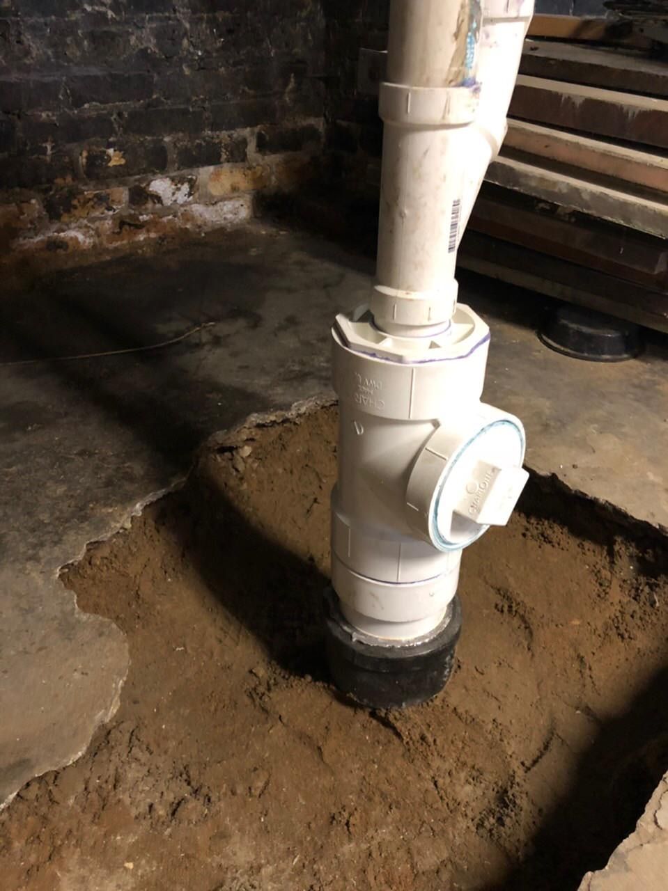 White PVC drain pipe in a dirt-filled square opening in a concrete floor, in a basement.