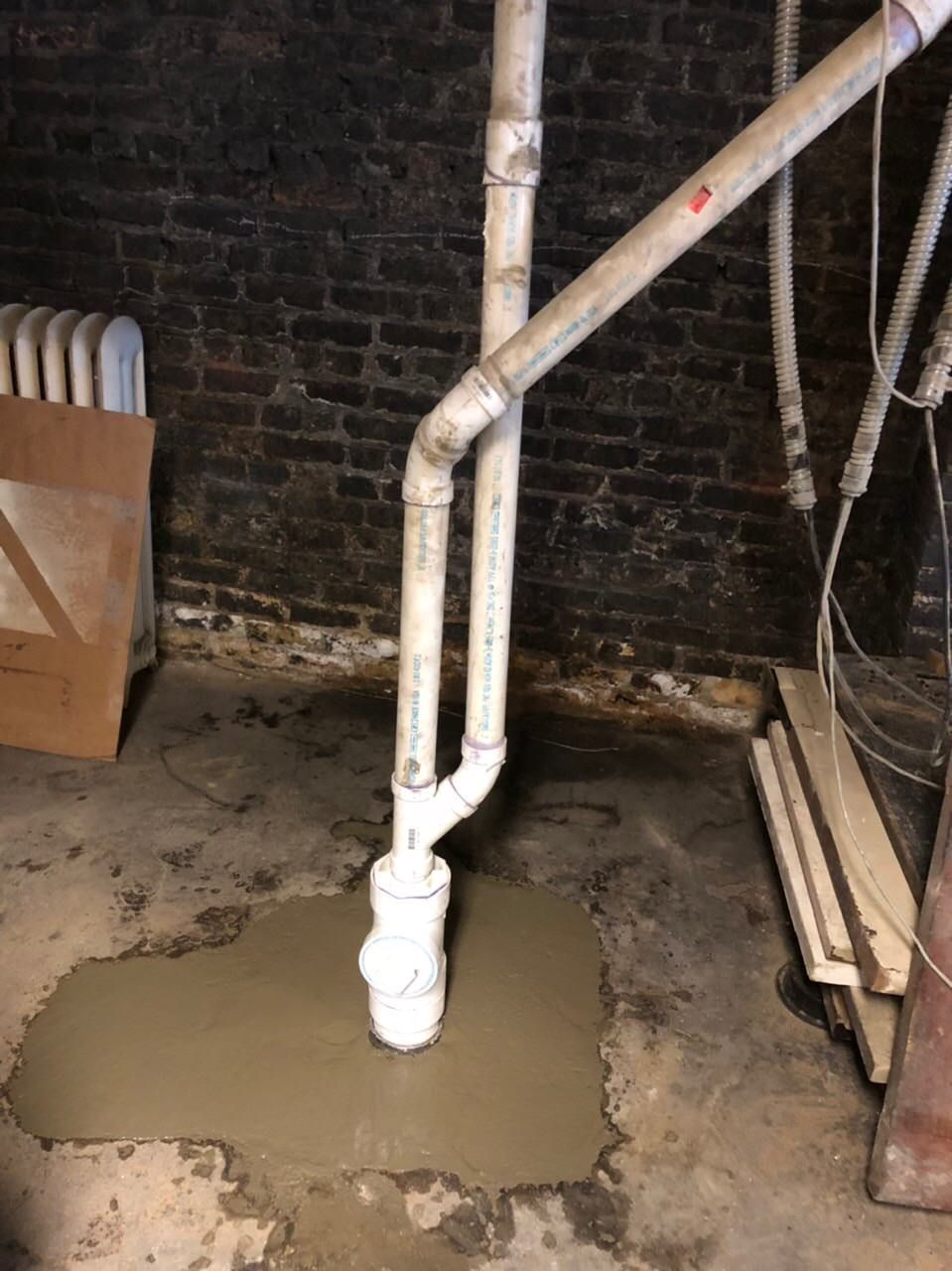 Basement sump pump installation: white pipes rise from a concrete-filled pit in front of a brick wall.