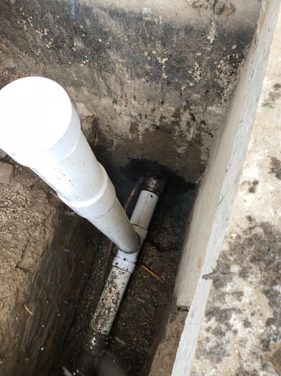 White PVC pipes in a trench, connecting to a drain, set in concrete, with dirt visible.