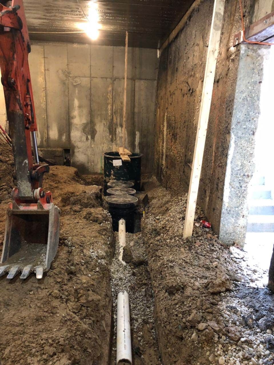 Trench with white pipe being installed, excavator, concrete wall, and dark septic tank.