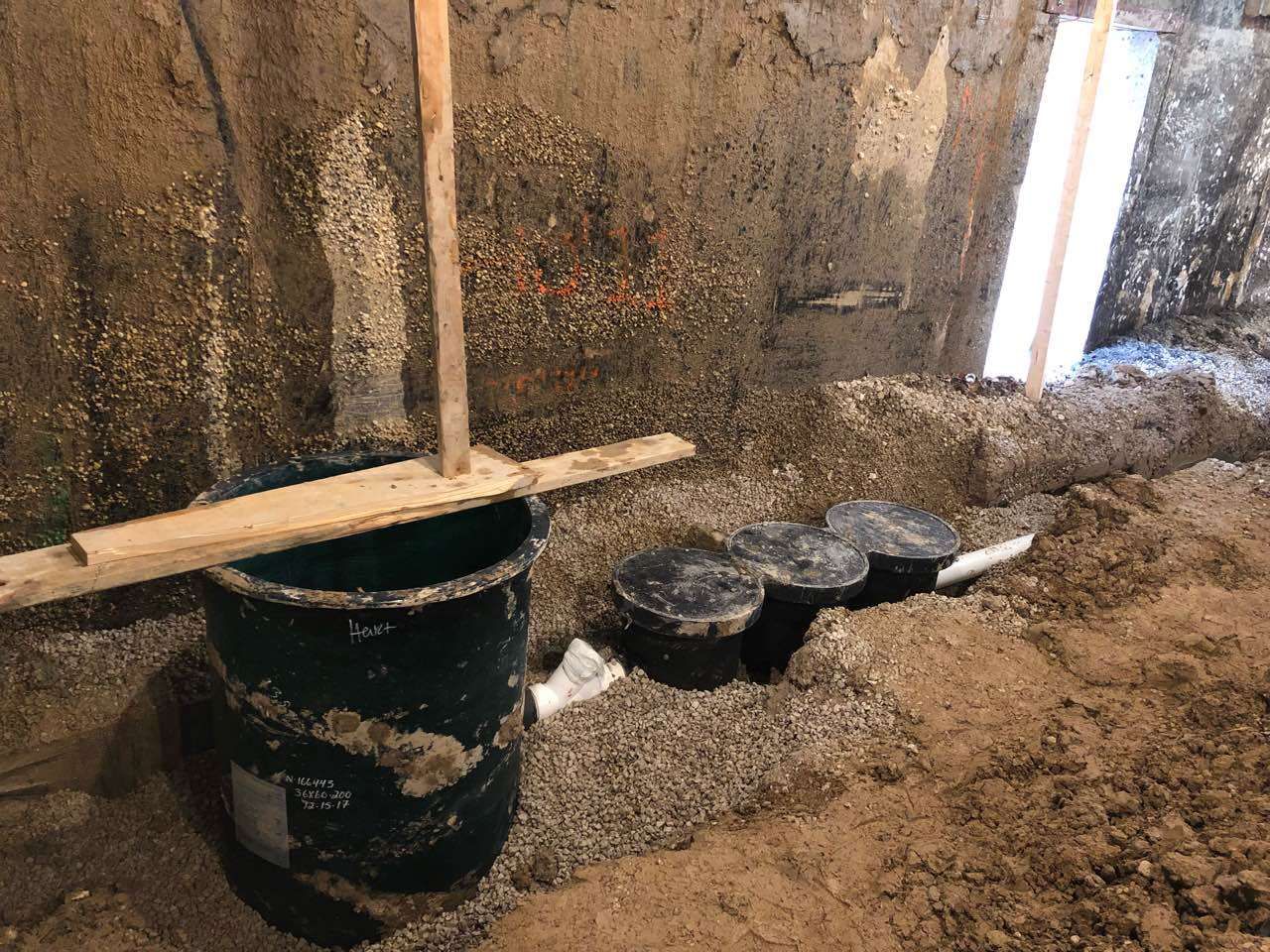 Dark green barrel with wood planks, three containers, and gravel on earthen floor, near a wall.