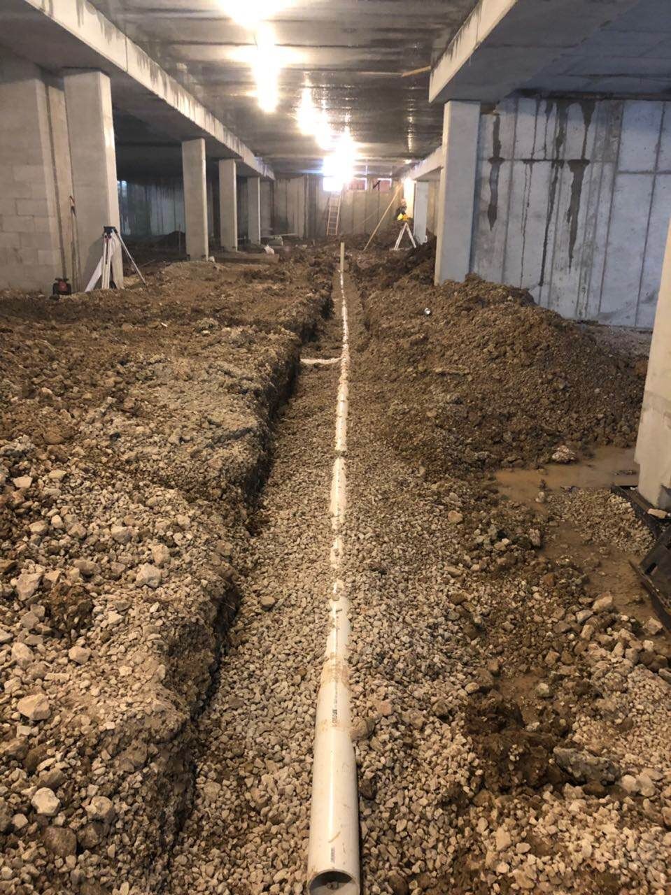 Construction site: A trench with white pipes, surrounded by gravel and dirt, under concrete beams.