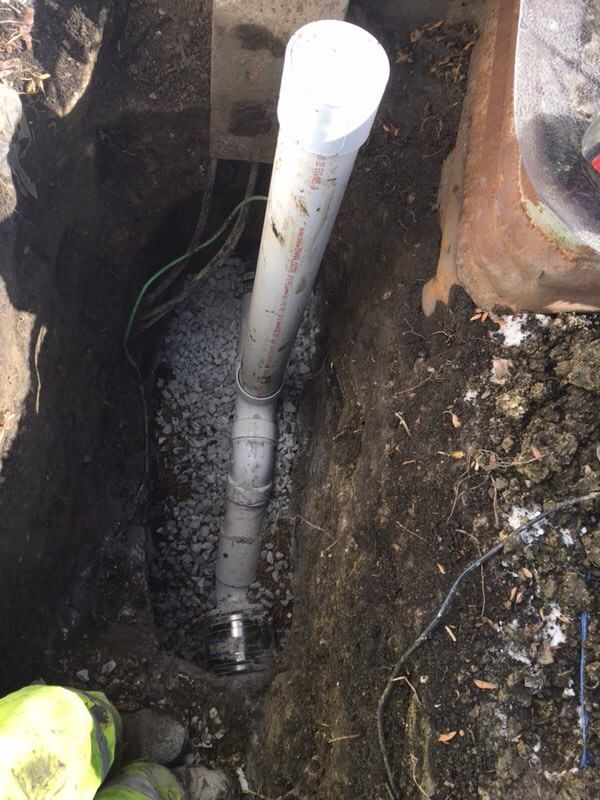 A vertical view of a pipe in a dug hole filled with gravel. Electrical wires and a concrete edge are visible.