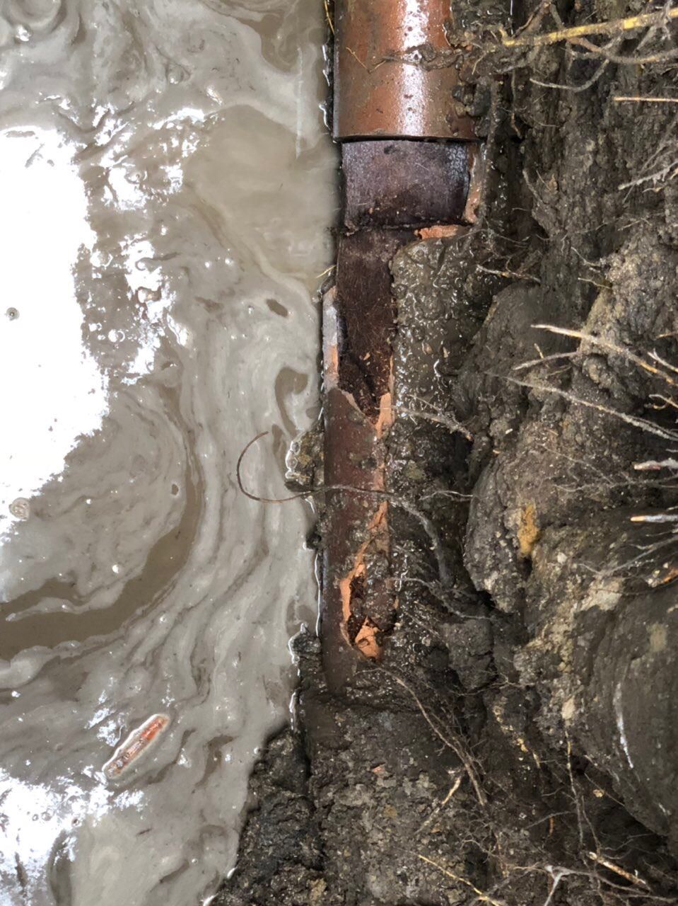 Damaged rusty pipe in a muddy trench with standing water. Brown dirt and brick visible.