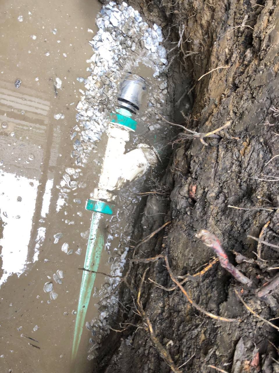 White PVC pipe with water spraying out, in a muddy trench.