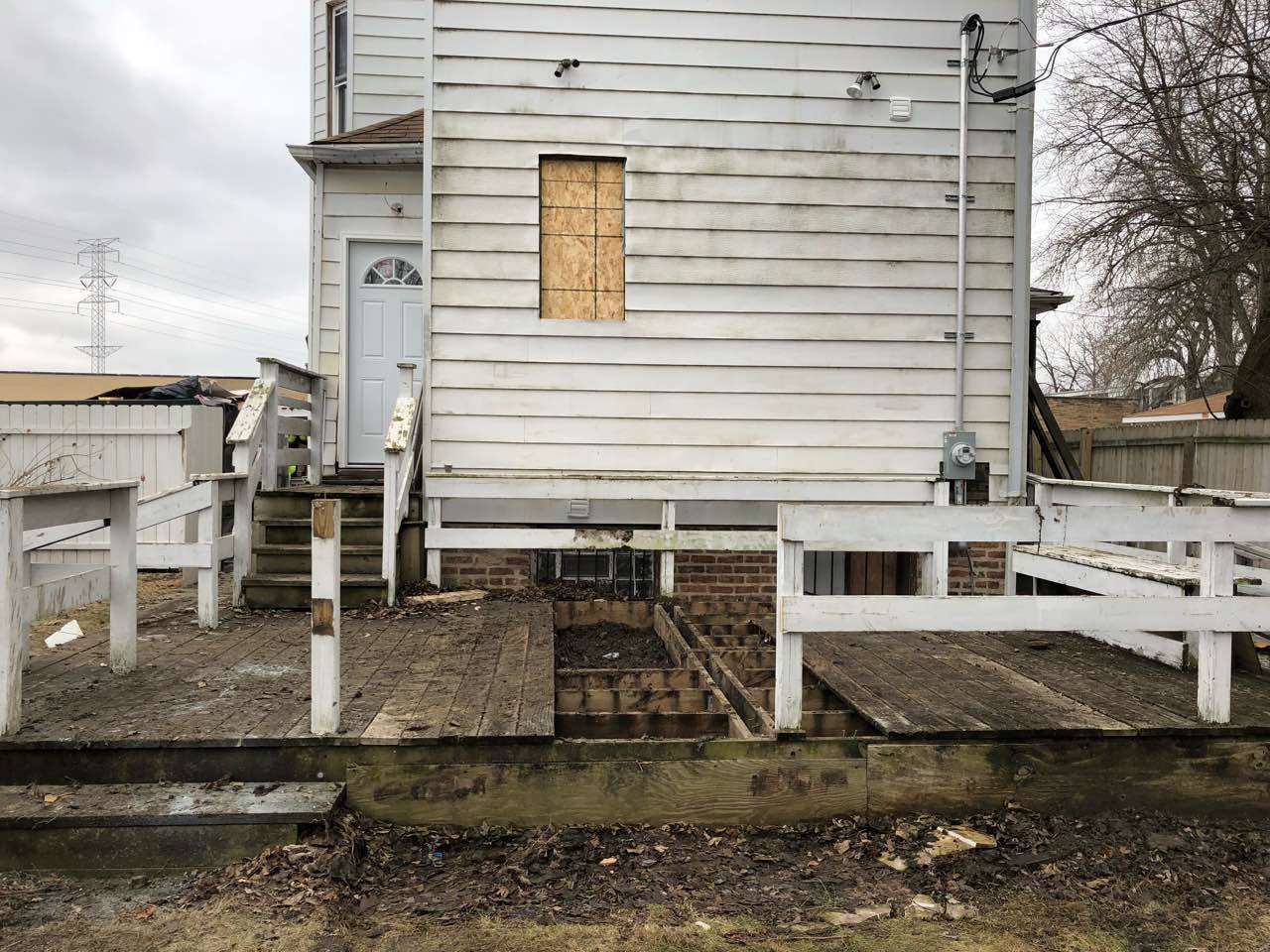 Dilapidated white house with a partially collapsed wooden deck, boarded-up window, and exposed foundation.