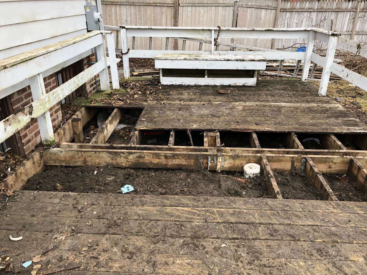 A damaged wooden deck with exposed joists, white railing, and debris.